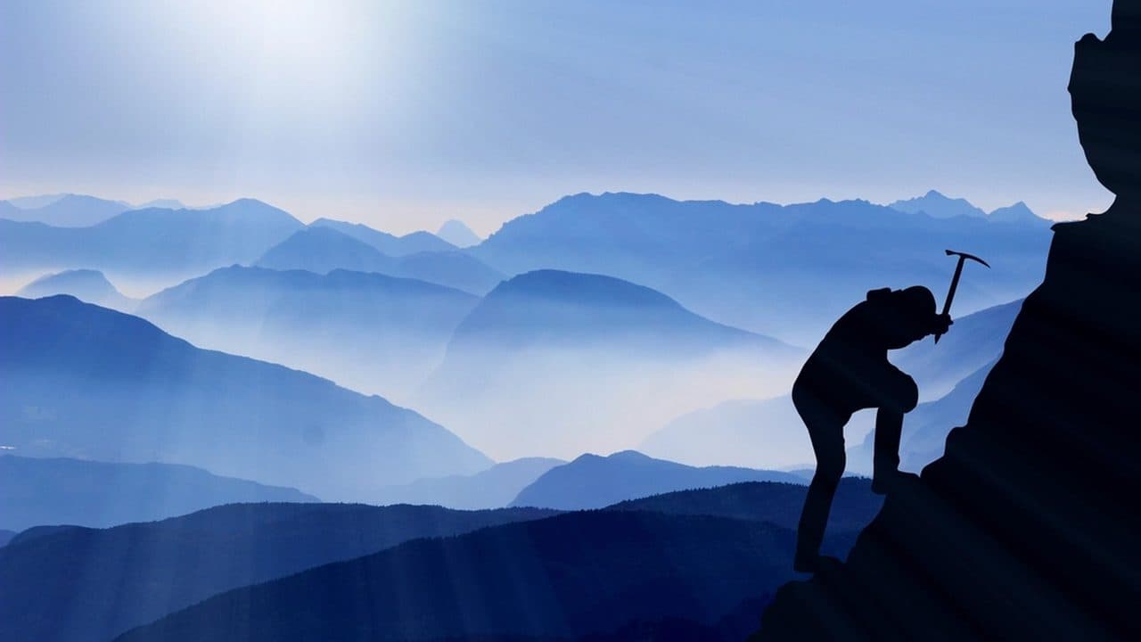 Silhouette of a climber with an ice axe pulling up a rock face, with hazy mountain ridges and a low sun behind